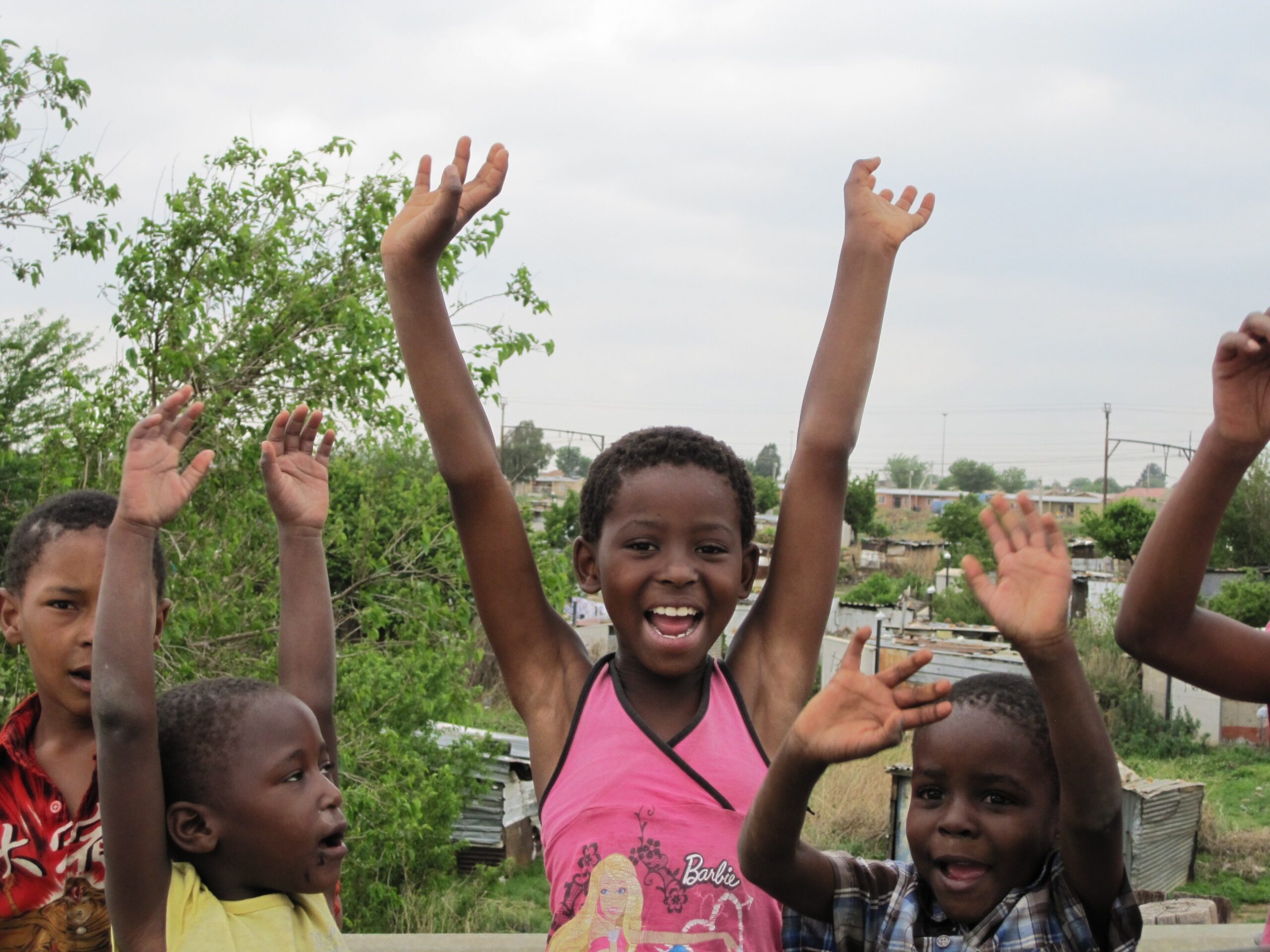 Children raising their hands and smiling in a community setting, representing youth wellbeing and mental health support in East Africa.